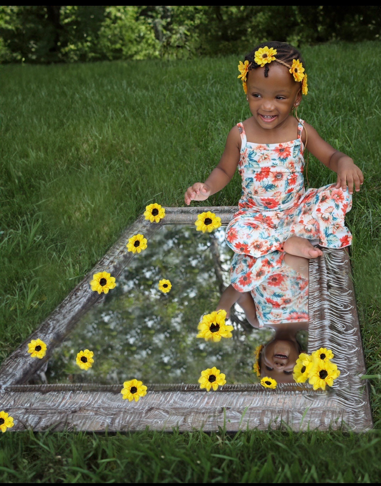 Child wearing flowers in her hair, seated beside a flower-covered mirror in the garden